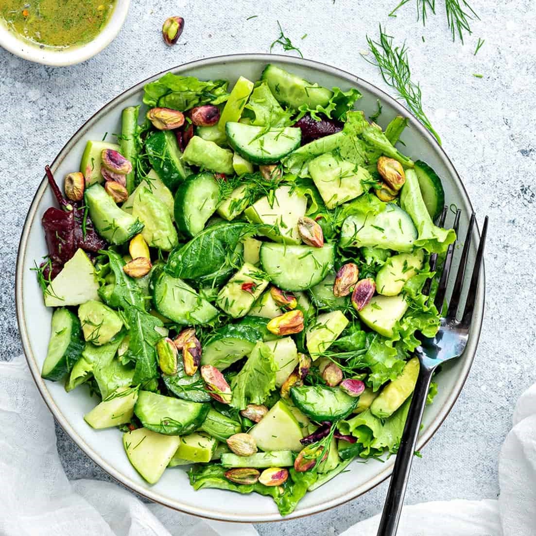 Overhead shot of a green salad that includes lettuce, spinach, cucumber, avocado, pistaschio and garnished with dill