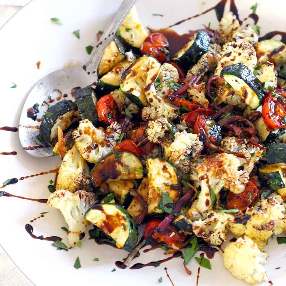 Close up shot of a plate of grilled veggies that includes courgettes, cauliflower florets, and tomatoes with a generous topping of balsamic glaze